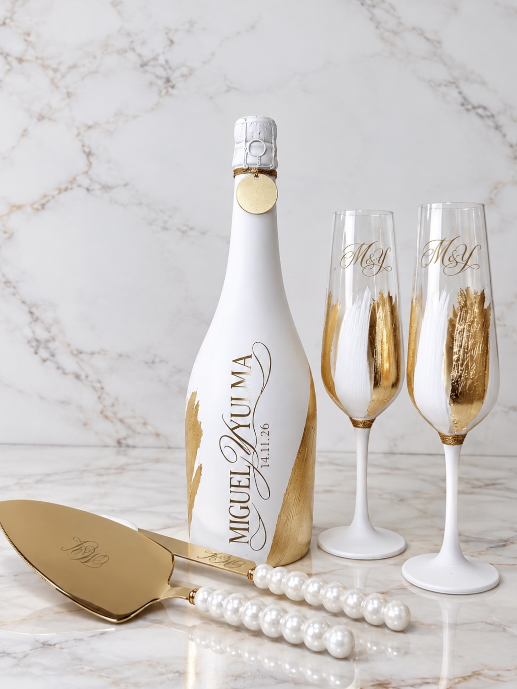 Botella y copas blancas con pan de oro y monograma personalizado junto a pala y cuchillo con perlas